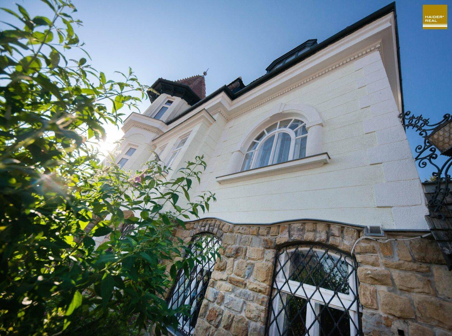 Historische Fassade mit Steinmauerwerk und großen Bogenfenstern, umgeben von üppigem Grün im Sonnenlicht.