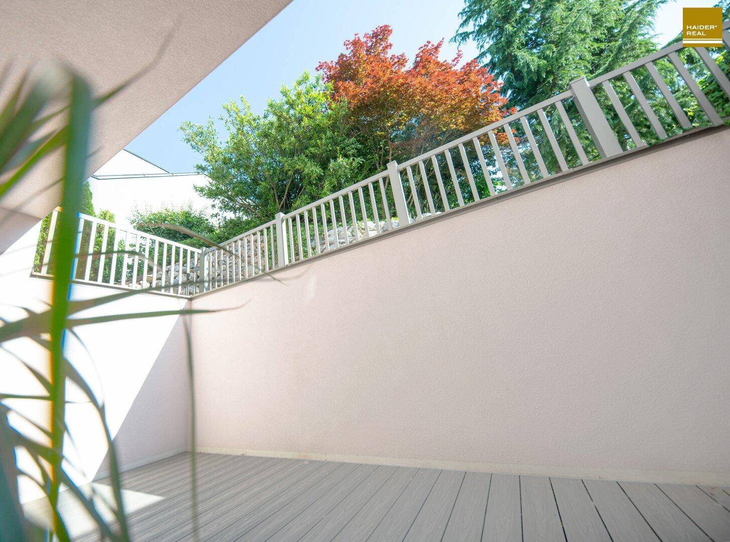 Heller Balkon mit Holzbelag und weißem Geländer, umgeben von grünen Bäumen.