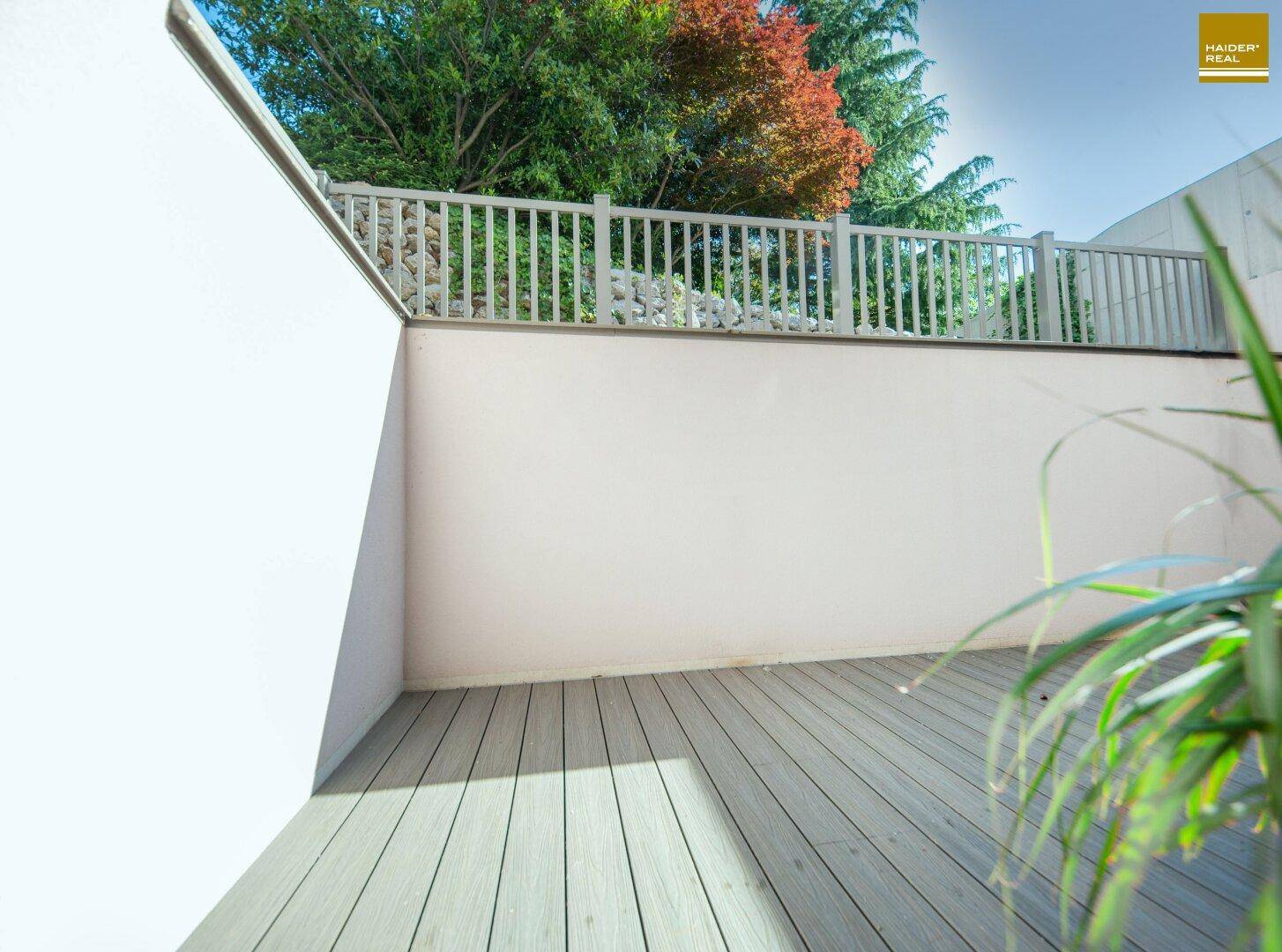 Heller Balkon mit Holzbelag und weißem Geländer, umgeben von grünen Pflanzen.