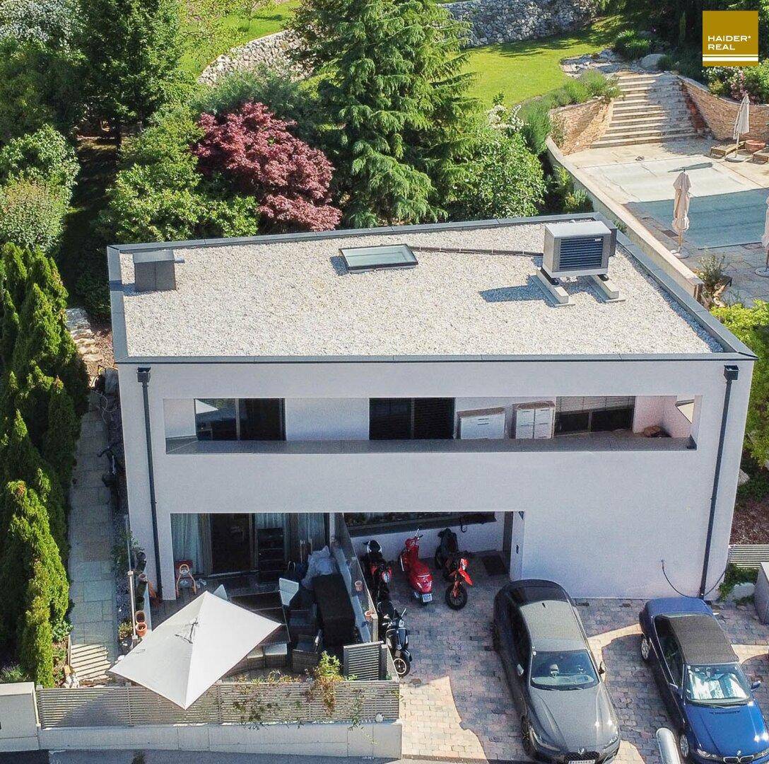 Luftaufnahme eines modernen Hauses mit Flachdach, Parkplätzen und üppiger grüner Umgebung.