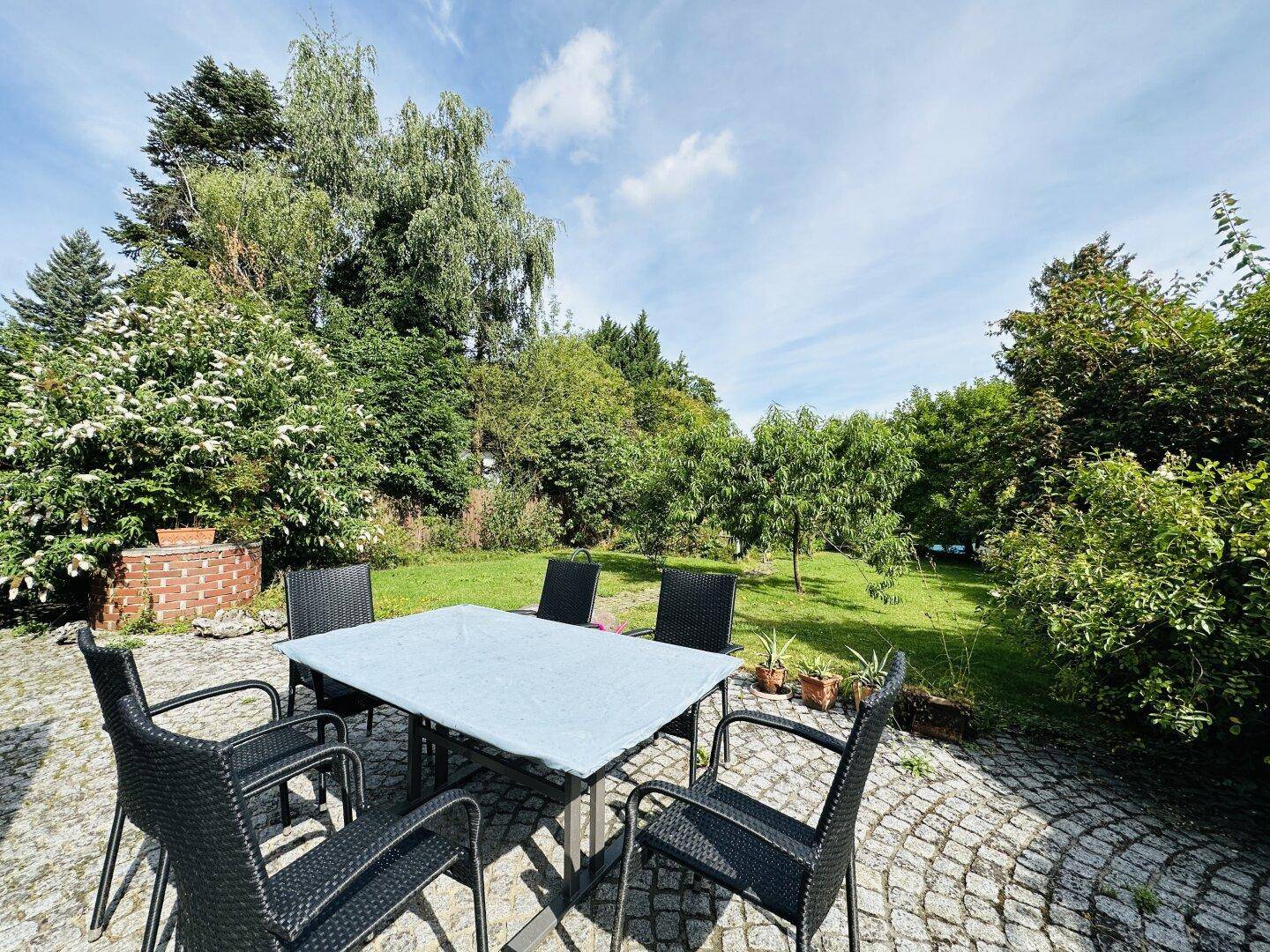 Gemütlicher Essbereich im Freien auf einer gepflasterten Terrasse, umgeben von einem grünen Garten.