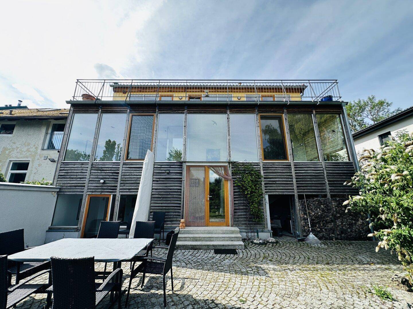 Rückseite eines modernen Hauses mit großer Glasfassade und einer gepflasterten Terrasse im Vordergrund.
