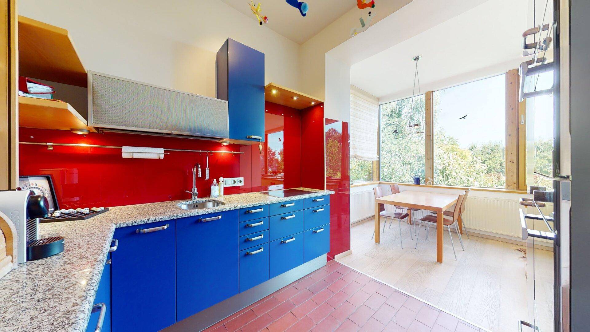 Moderne Küche mit blauen Schränken, roter Rückwand und einem Essbereich mit Blick ins Grüne.