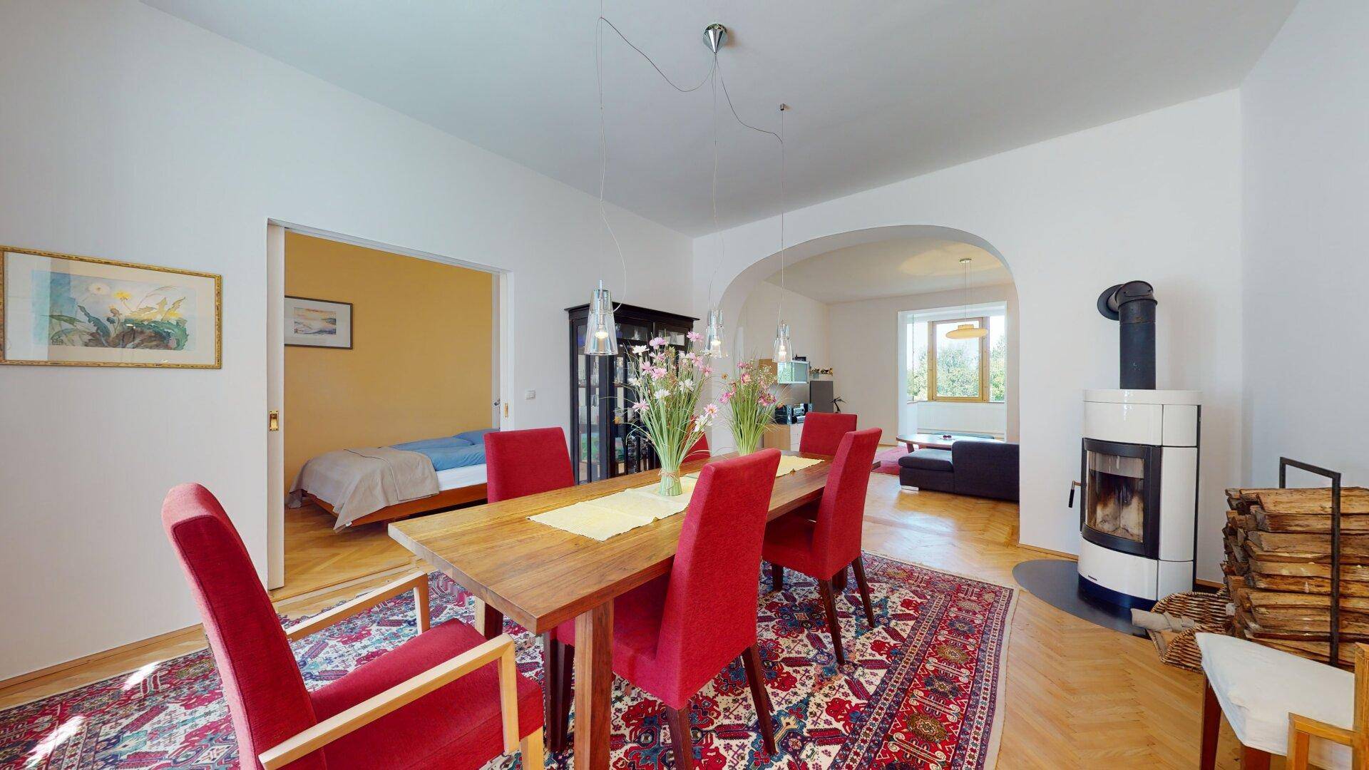 Offener Wohn- und Essbereich mit Kaminofen, Holztisch und Zugang zu einem Schlafzimmer.