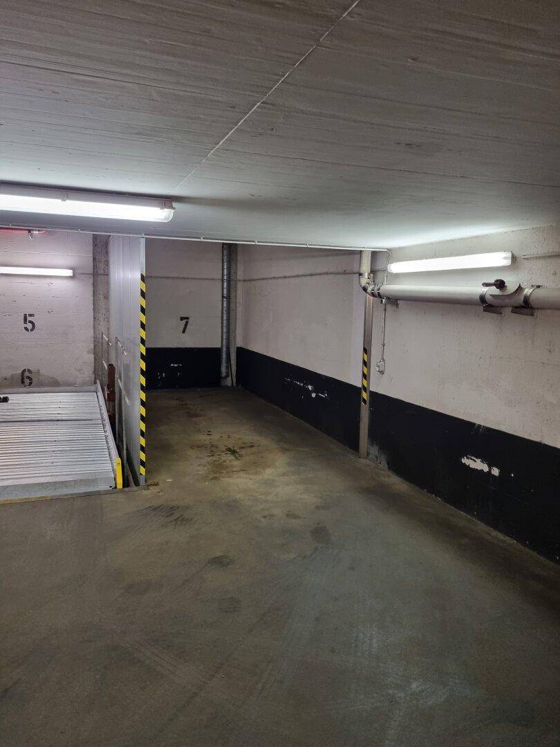 Gang in der Tiefgarage mit Betonboden, sichtbaren Rohren und Leuchtstoffröhrenbeleuchtung.