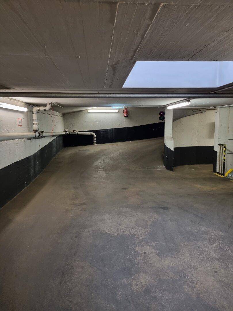 Tiefgaragenrampe mit Betonoberflächen und Leuchtstoffröhrenbeleuchtung, die zu weiteren Parkebenen führt.