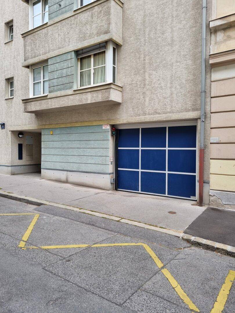 Fassade eines Gebäudes mit einer blauen Garagentür auf Straßenniveau und gelben Markierungen auf der Fahrbahn.