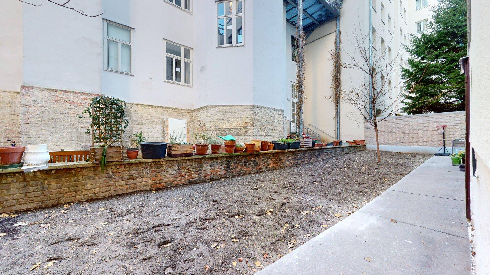 Länglicher Hinterhofgarten mit Ziegelmauer und Topfpflanzen, umgeben von Wohngebäuden.