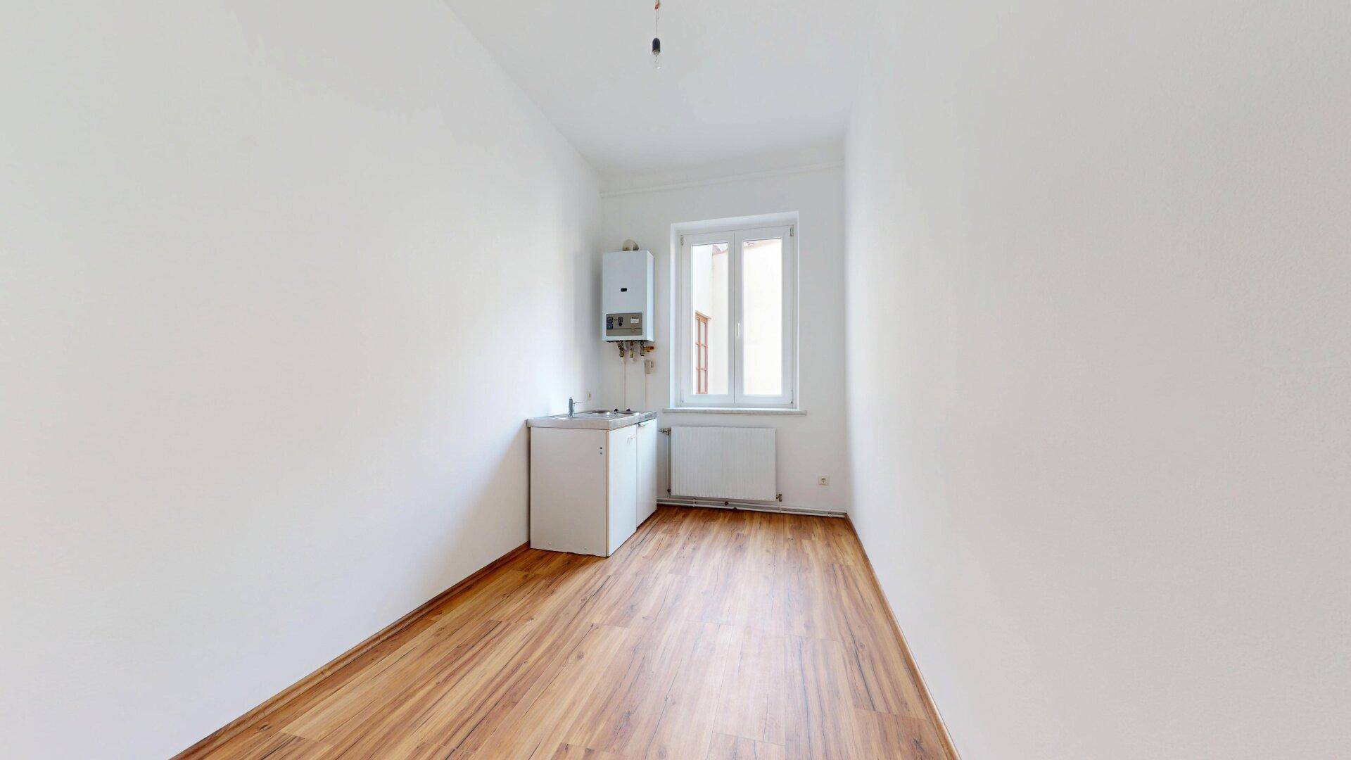 Leerer Küchenbereich mit Fenster, Gastherme und kleinem Unterschrank, bereit zur Einrichtung.
