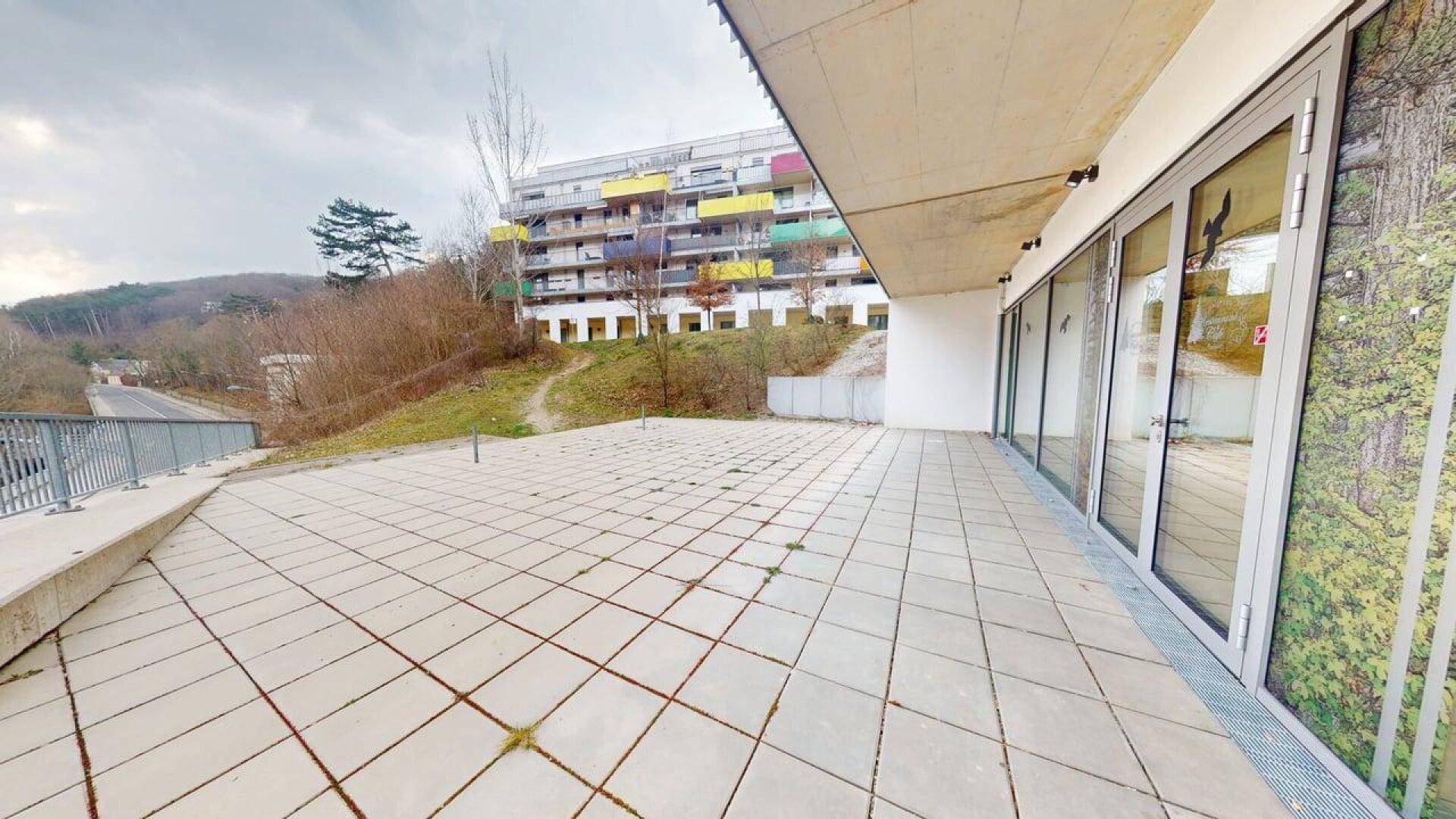 Weitläufige Außenterrasse mit Fliesenboden und Blick auf ein modernes Gebäude und die Landschaft.