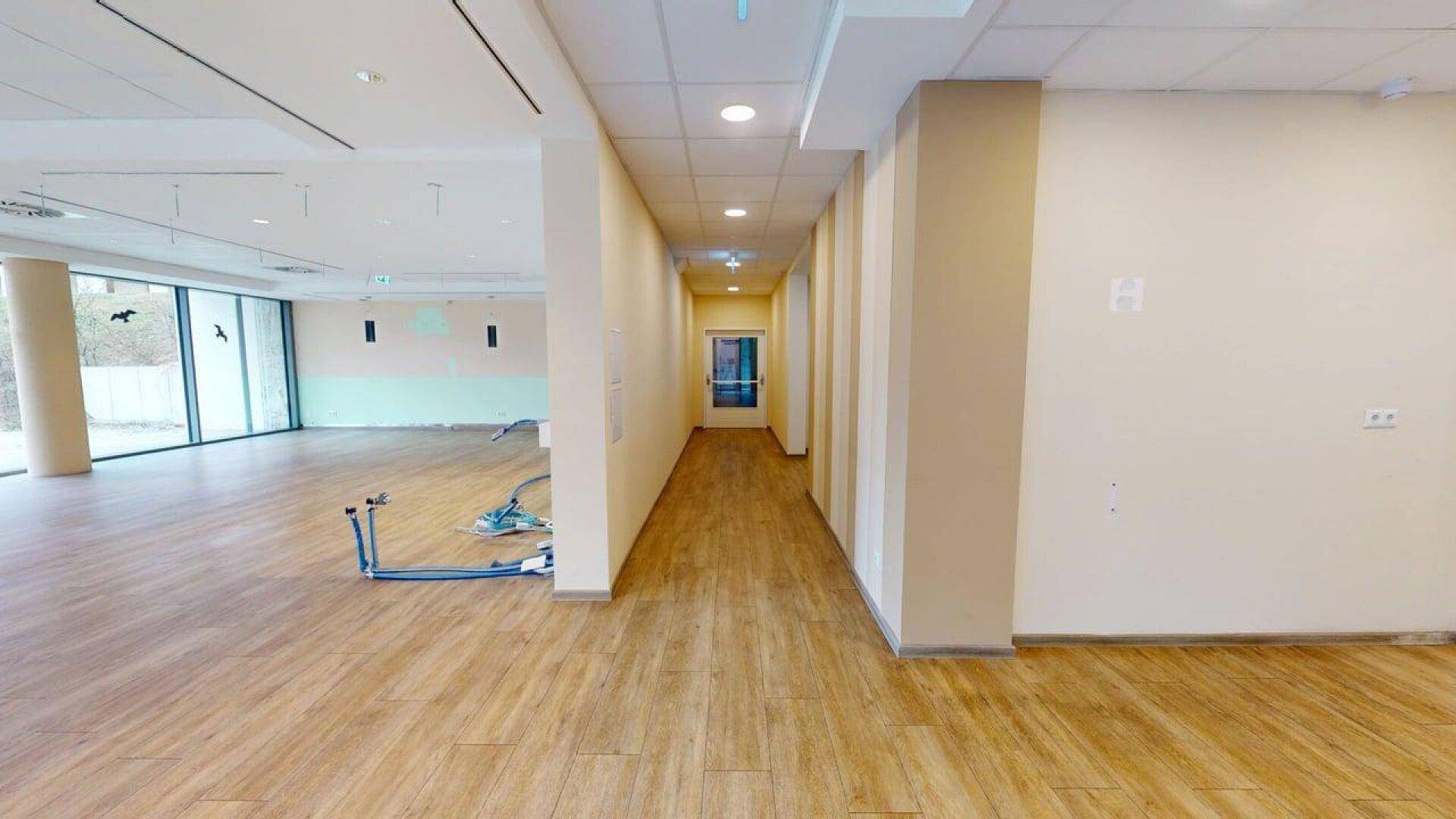 Heller Flur in einem Gewerberaum mit Holzboden, der zu einem offenen Bereich mit großen Fenstern führt.