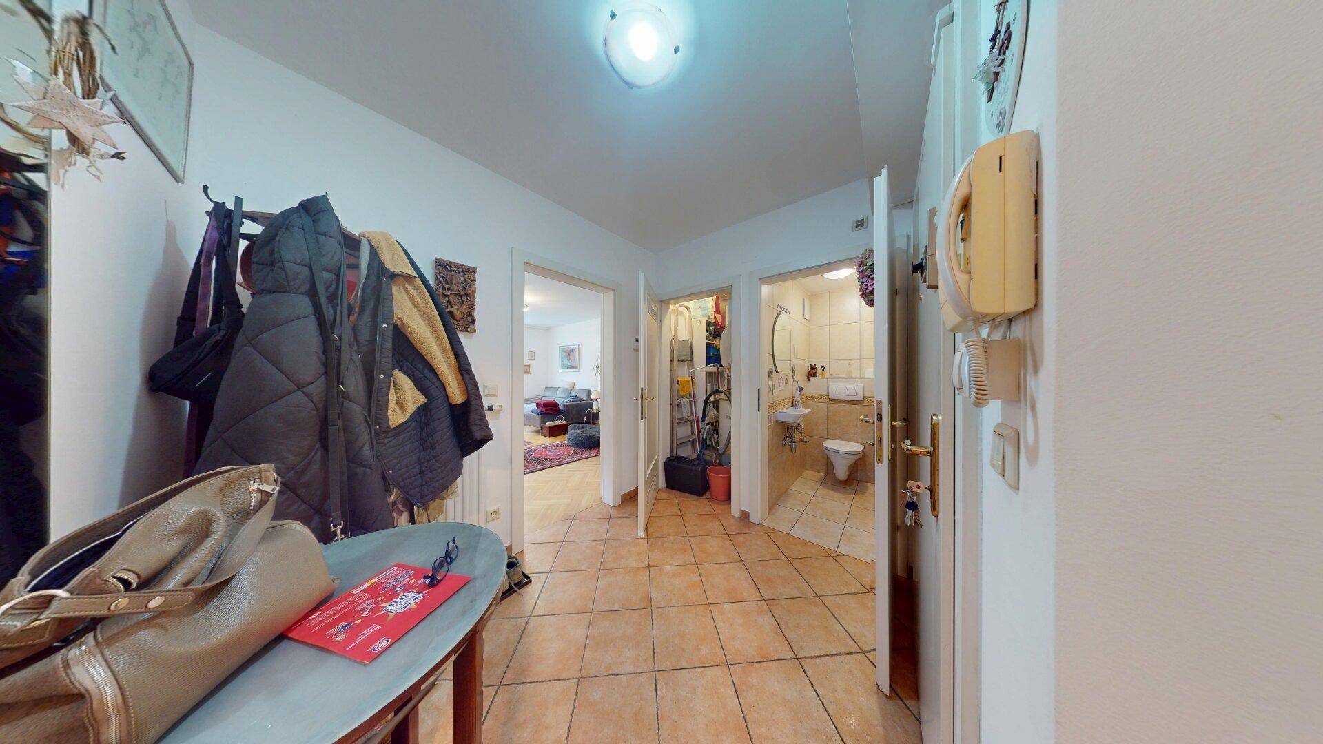 Flur mit beigefarbenen Fliesen, Gegensprechanlage und Blick in das Badezimmer.