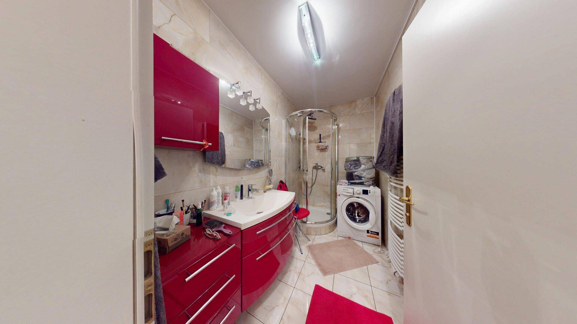Modern gefliestes Badezimmer mit Duschkabine, Waschmaschine und rotem Waschtisch.