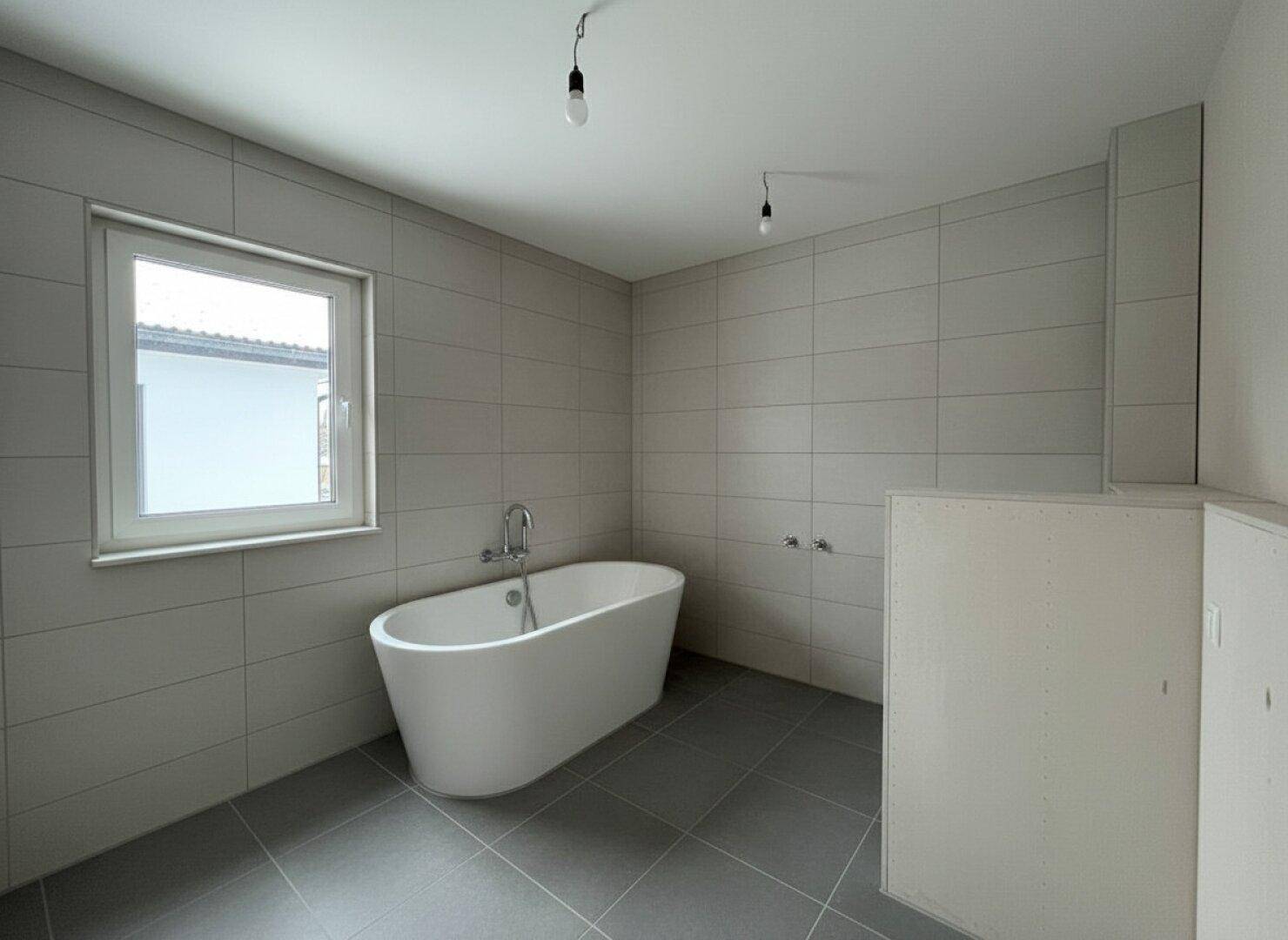 Modernes Badezimmer mit freistehender Badewanne, grauen Fliesen und Fenster.
