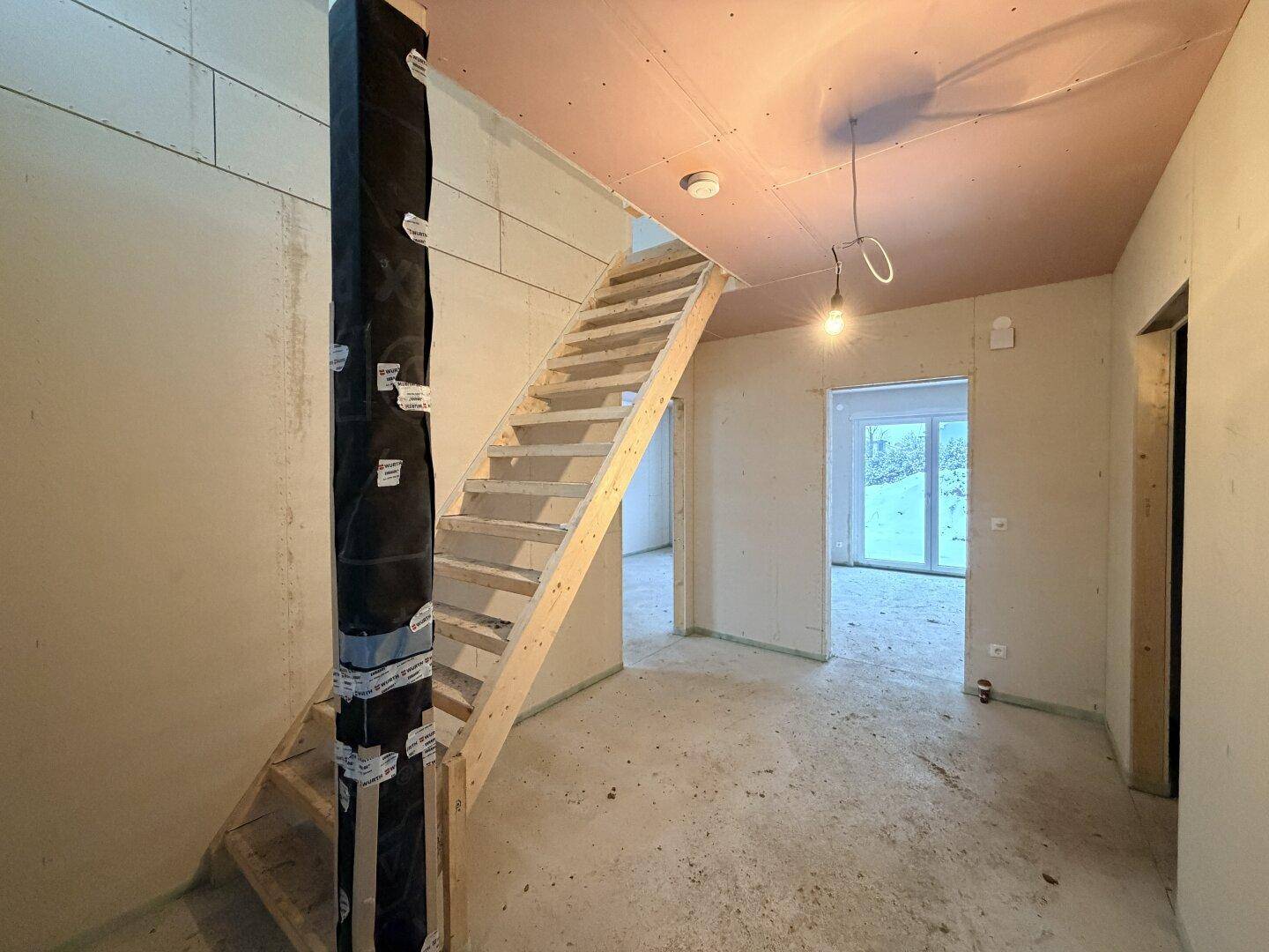 Unfertiger Flur im Rohbau mit Holztreppe und Blick in weitere Räume.