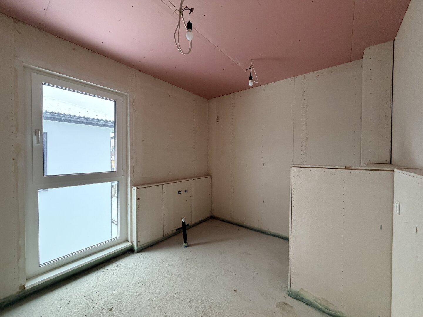 Unfertiges Badezimmer im Rohbau mit Fenster und vorbereiteten Anschlüssen für eine Badewanne.