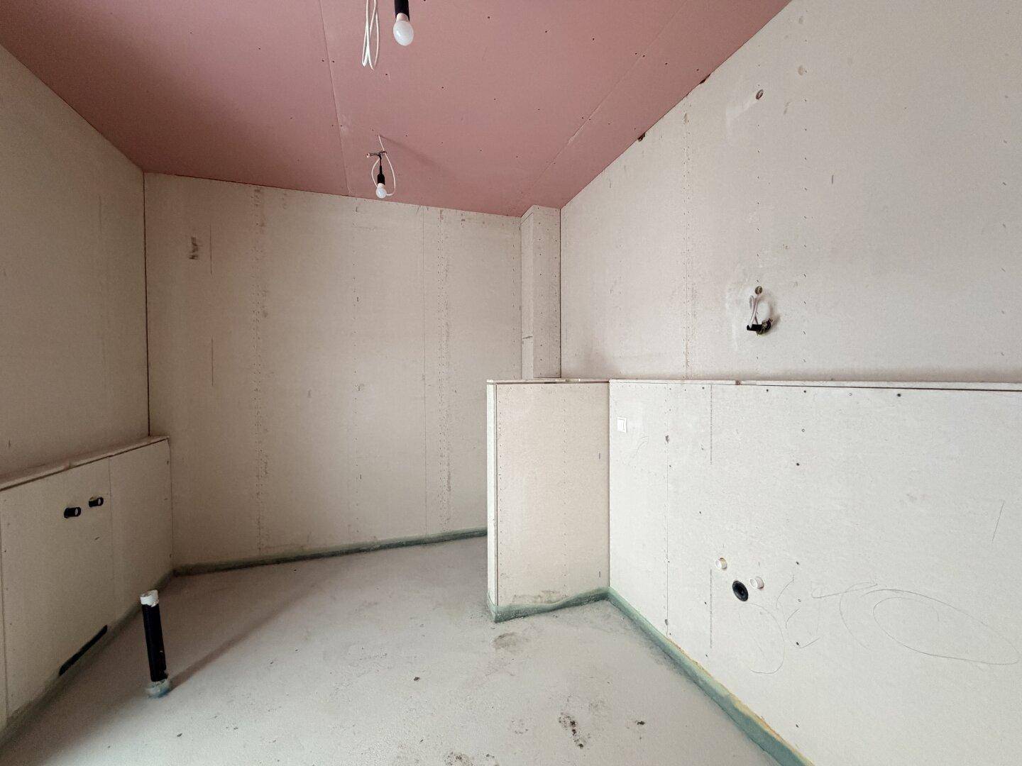 Unfertiges Badezimmer im Rohbau mit vorbereiteten Anschlüssen und rosa Decke.