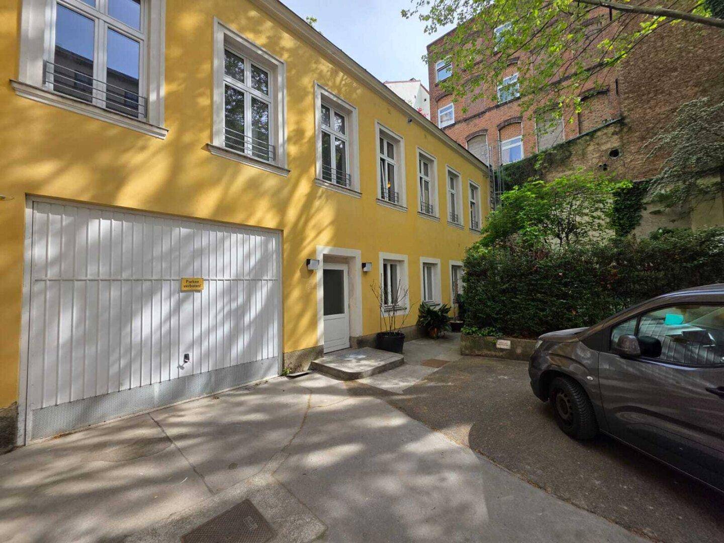 Gelbes Gebäude mit weißem Garagentor und Eingangstür, umgeben von Bäumen und einem gepflasterten Hof.
