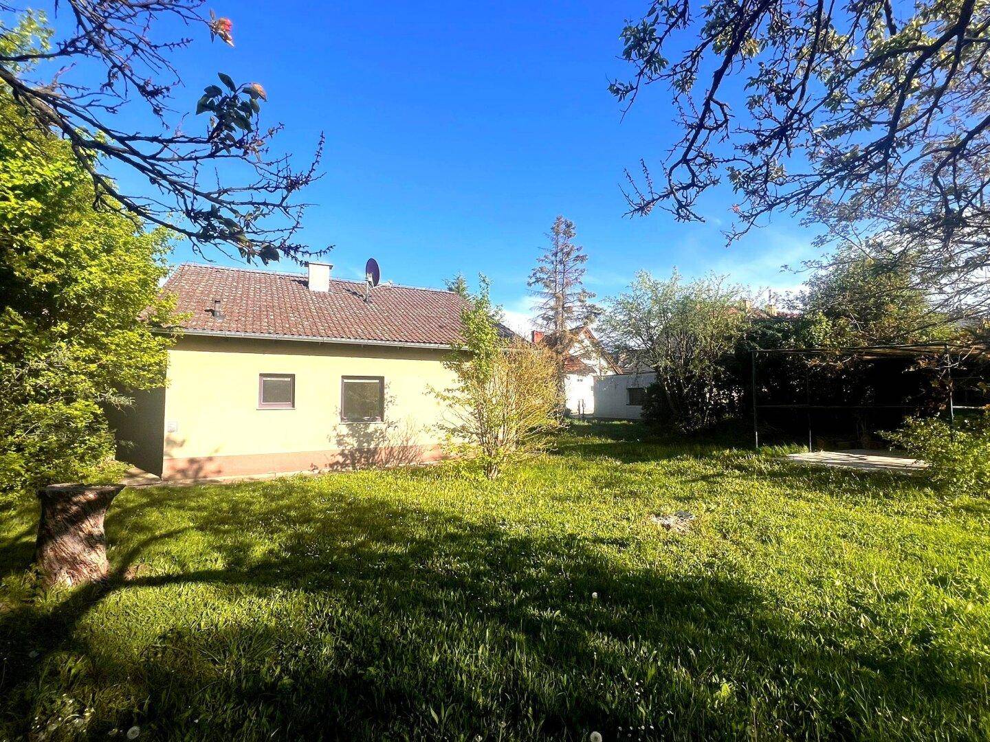Seitenansicht des Hauses mit Ziegeldach, umgeben von einem sonnigen Garten und Bäumen.