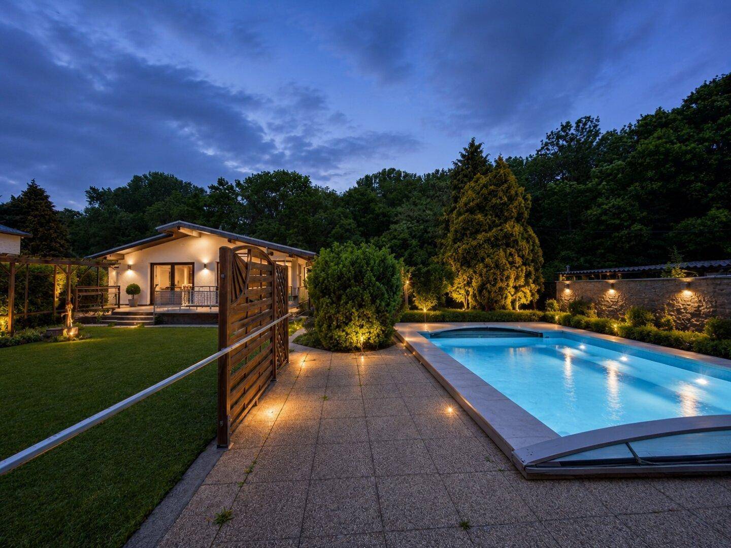Beleuchteter Poolbereich und renoviertes Haus bei Nacht mit atmosphärischer Gartenbeleuchtung.