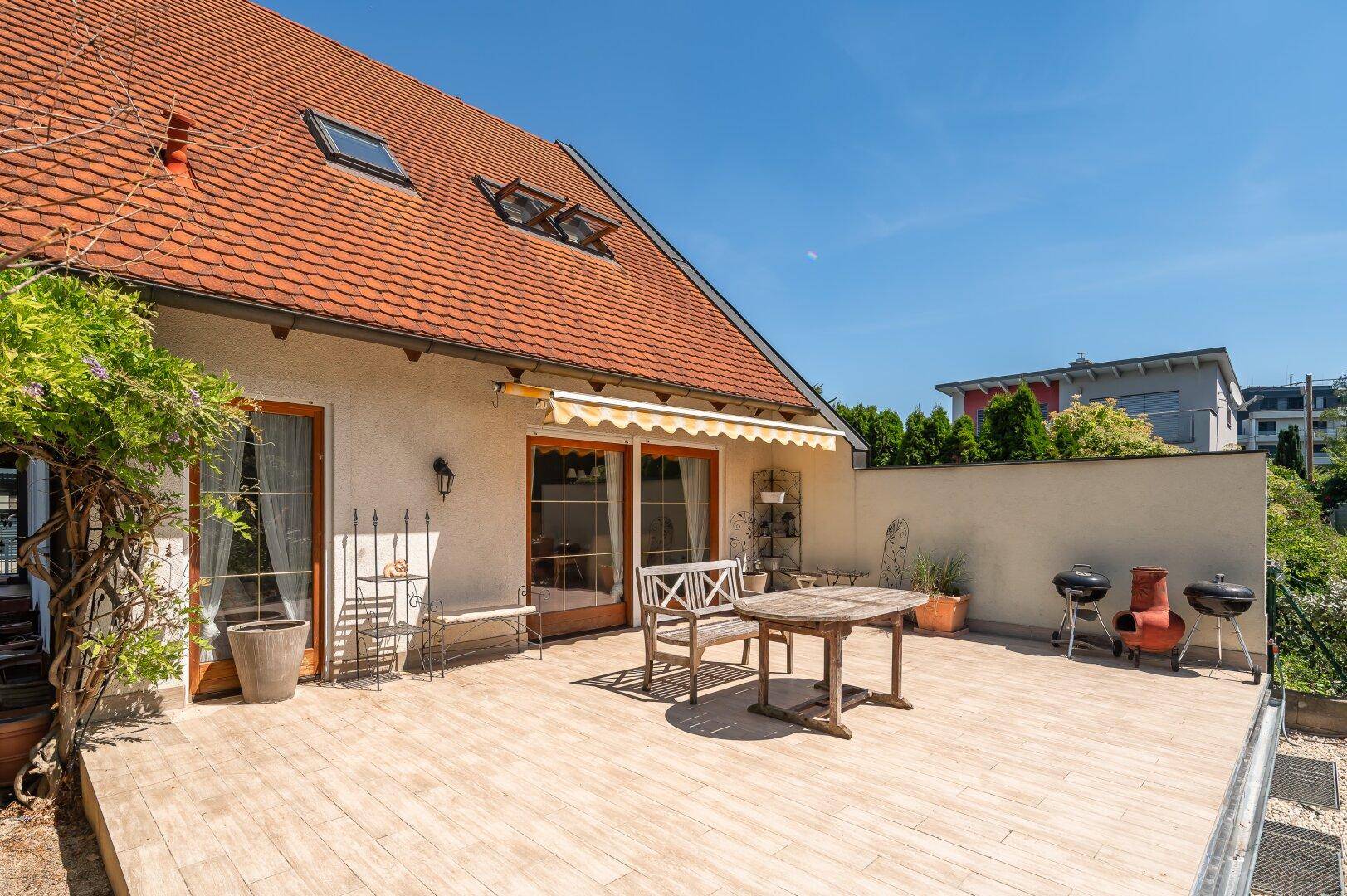 Große Sonnenterrasse am Haus mit Markise, Gartenmöbeln und Grillbereich unter einem Ziegeldach.