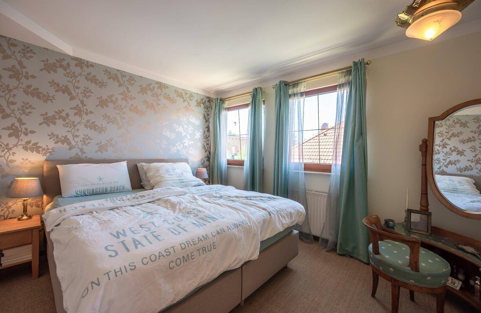 Gemütliches Schlafzimmer mit Doppelbett, gemusterter Tapete und zwei Fenstern mit blauen Vorhängen.