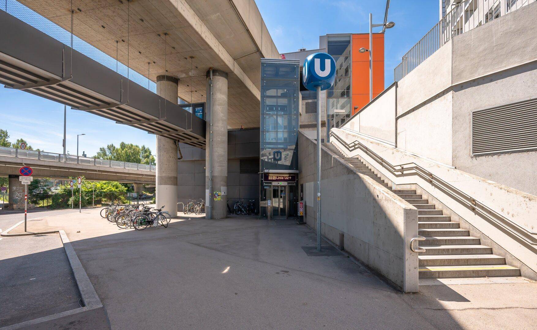 Moderner U-Bahn-Eingang mit Aufzug, Treppen und Fahrradabstellplätzen unter einer Hochbahnbrücke.