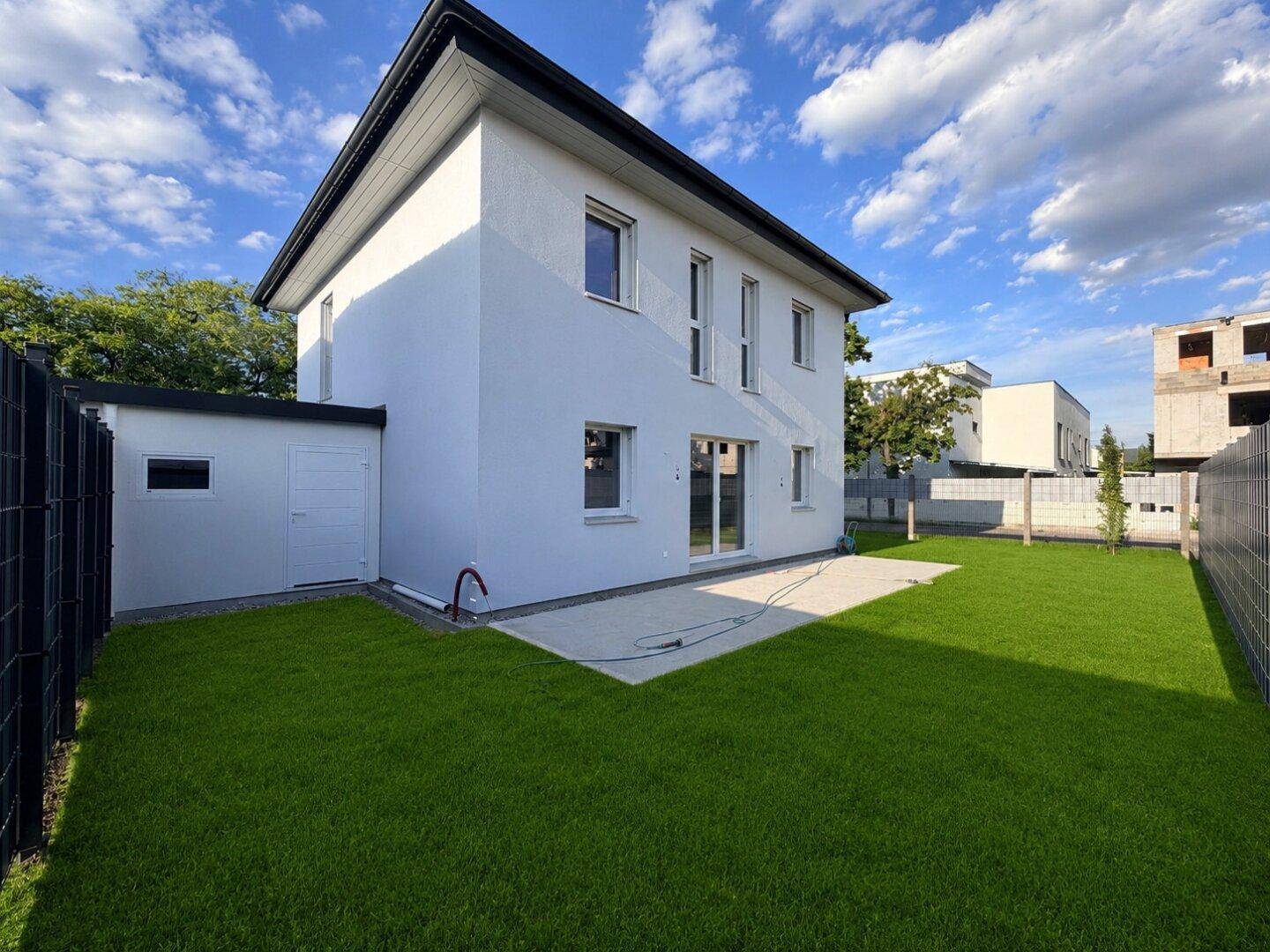 Hinterhof eines Neubaus mit weitläufigem Rasen, kleiner Terrasse und einem Gartenschuppen.