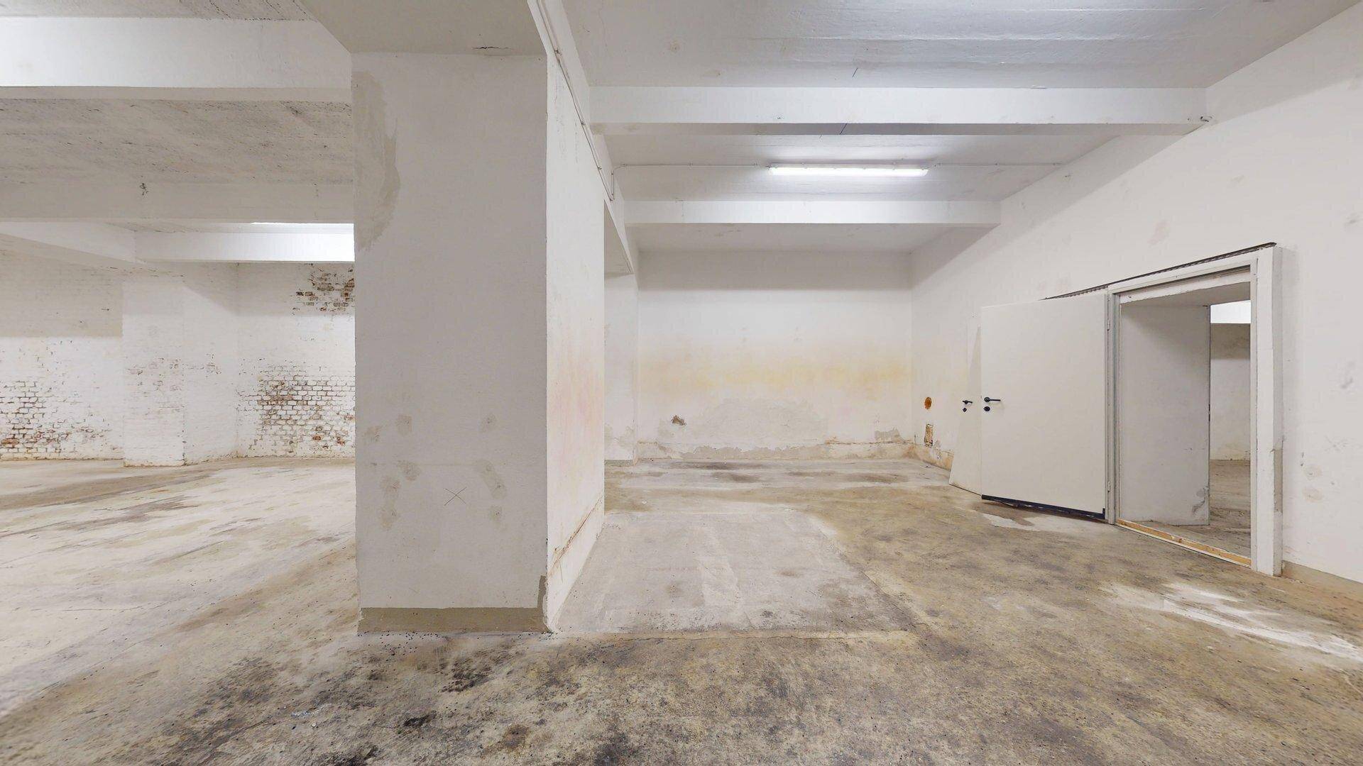 Weitläufiger Kellerbereich mit Betonboden, weiß gestrichenen Ziegelwänden und offenem Zugang zu weiteren Räumen.