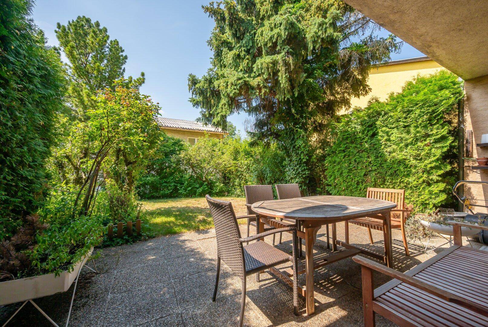 Sonniger Terrassenbereich mit Holztisch und Stühlen, umgeben von üppigem Grün.