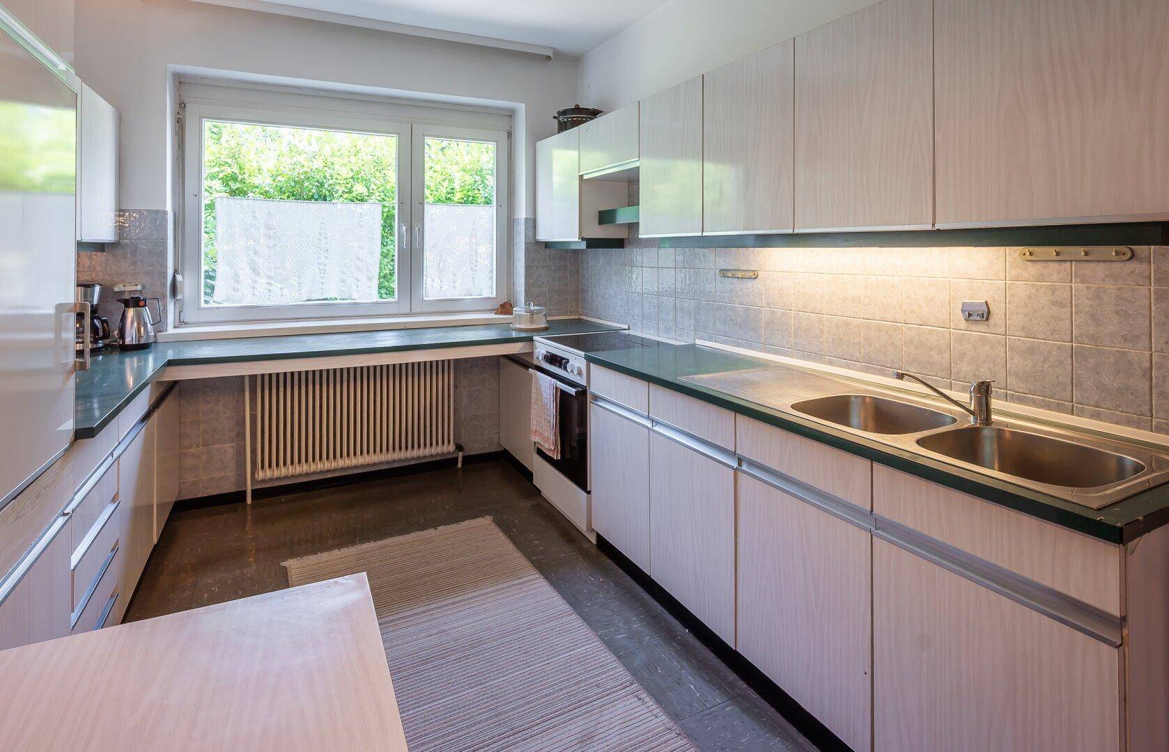Funktionale Küche mit hellen Schränken, grünen Arbeitsplatten und Fensterblick ins Grüne.
