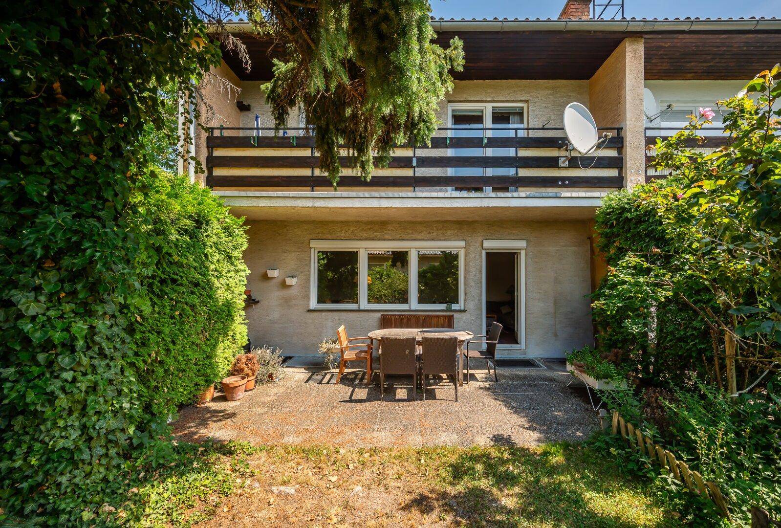 Terrasse mit Gartenmöbeln und Balkon darüber, umgeben von viel Grün.