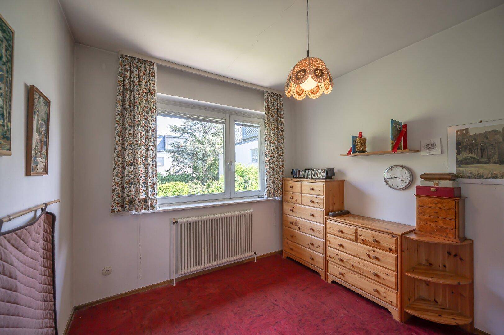 Helles Schlafzimmer mit rotem Teppichboden und Holzkommoden unter dem Fenster.