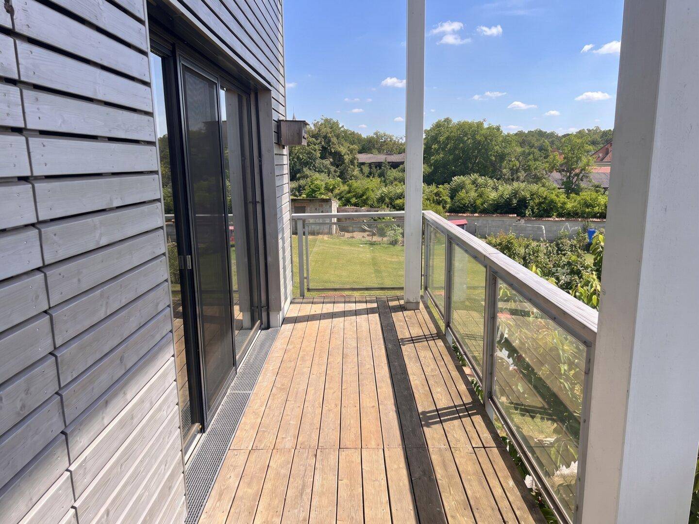 Geräumiger Balkon mit Holzboden und Glasgeländer, bietet einen weiten Blick ins Grüne.