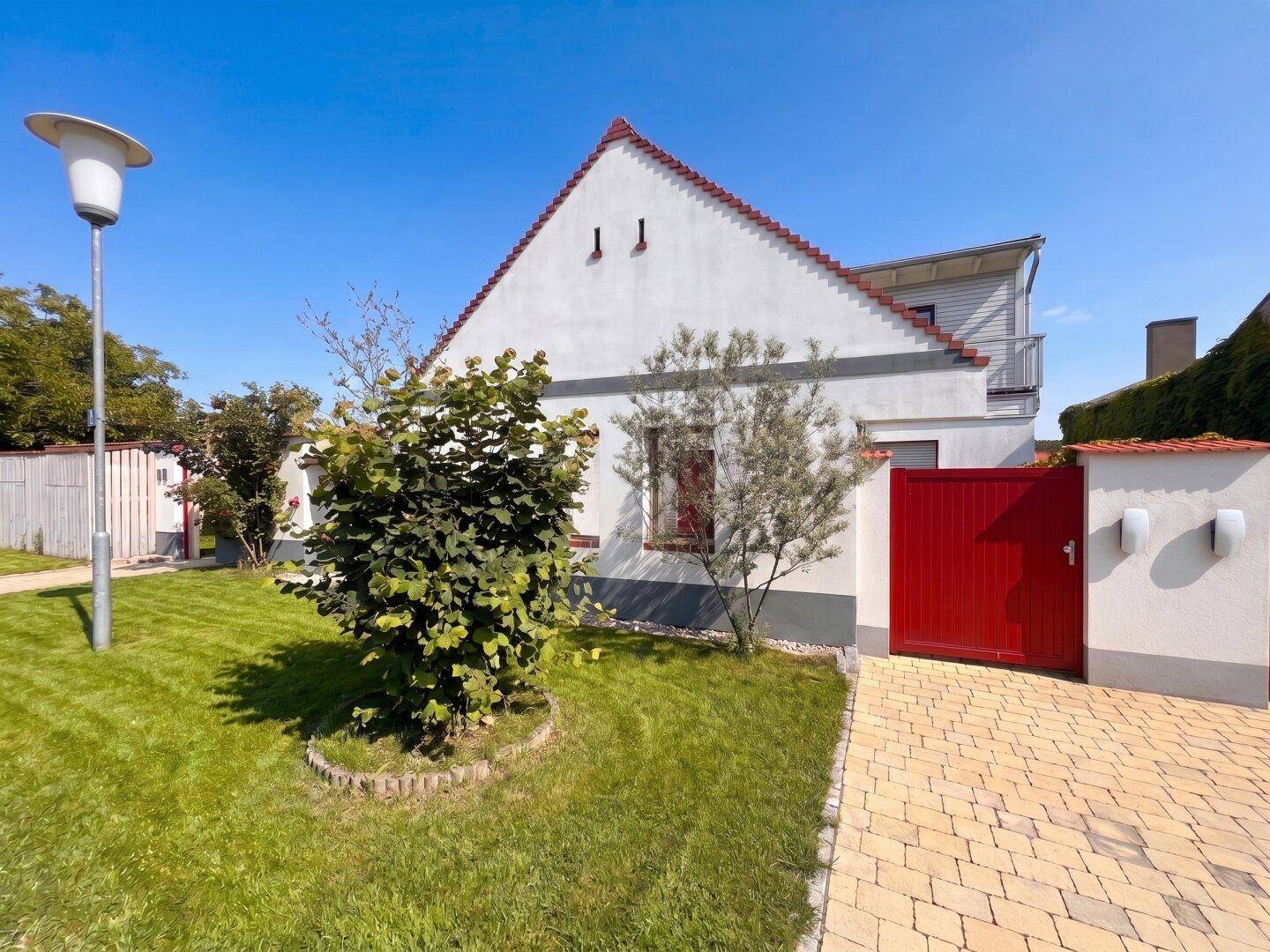 Ansicht des Hauses von vorne mit gepflegtem Rasen, Bäumen und rotem Eingangstor.