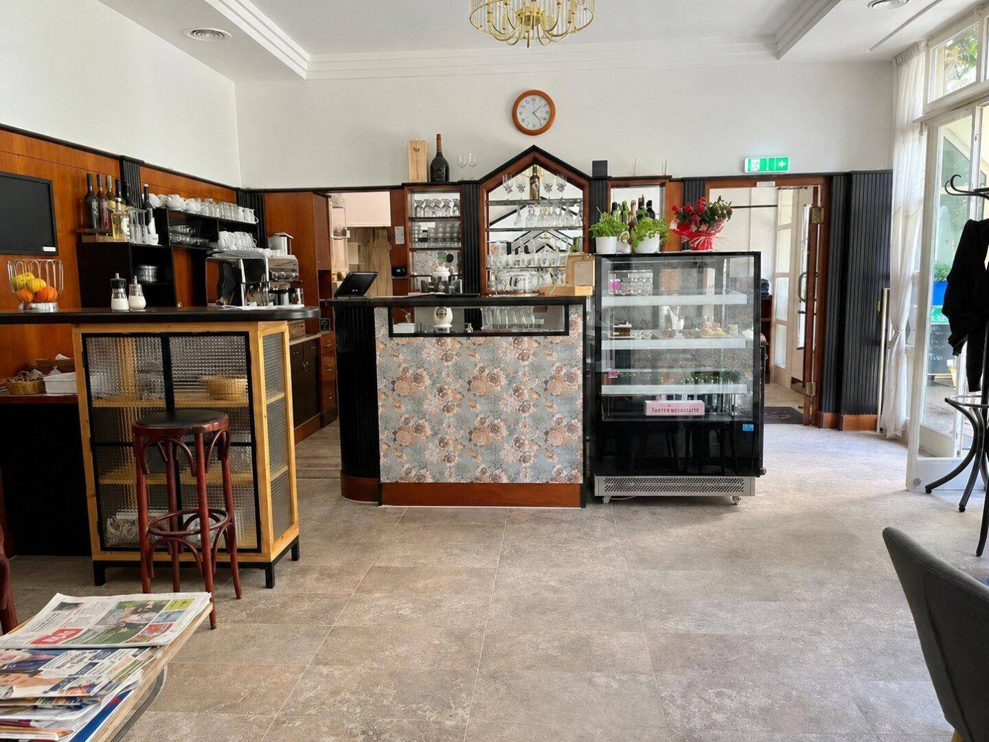 Weitläufiger Café-Bereich mit Theke, Kuchenvitrine und Sitzgelegenheiten, ergänzt durch eine Zeitungsablage.
