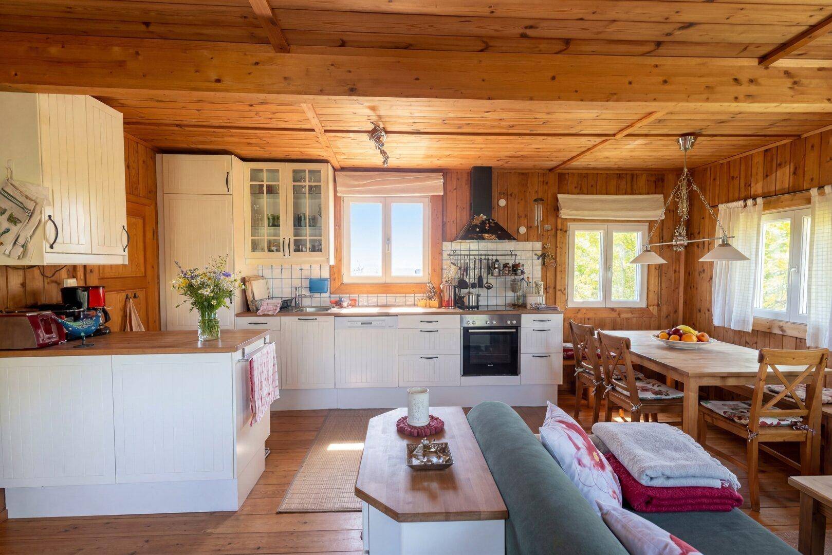 Offener Wohn-Essbereich mit Holzwänden und -decke, weißer Einbauküche und Esstisch.