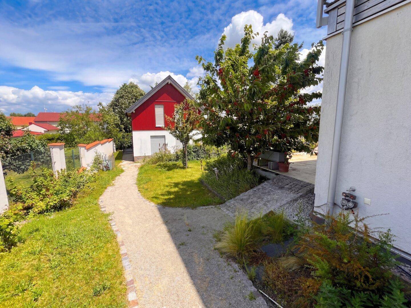 Gartenweg führt durch die grüne Anlage zum roten Haus unter blauem Himmel.