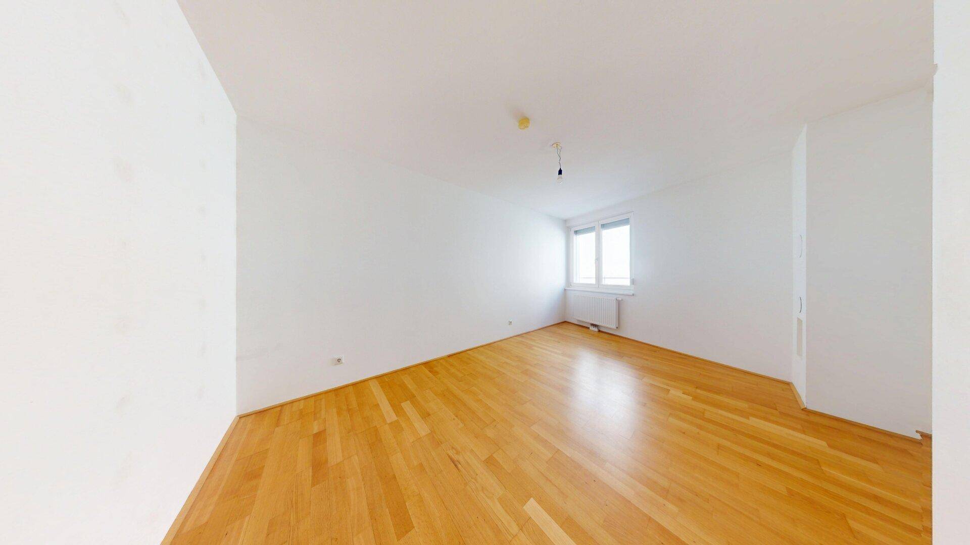 Leeres Schlafzimmer mit hellem Parkettboden und großem Fenster.