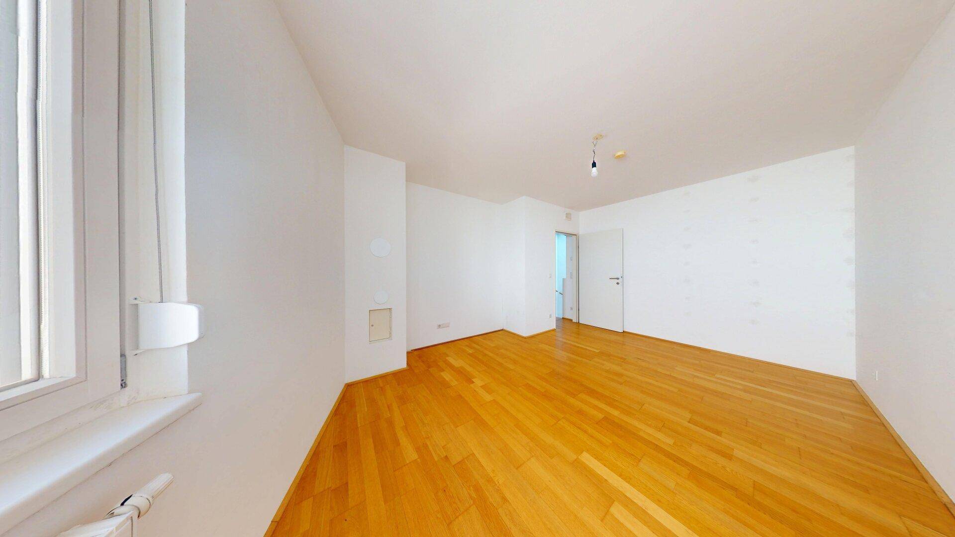 Geräumiges, leeres Zimmer mit Parkettboden und Fenster.