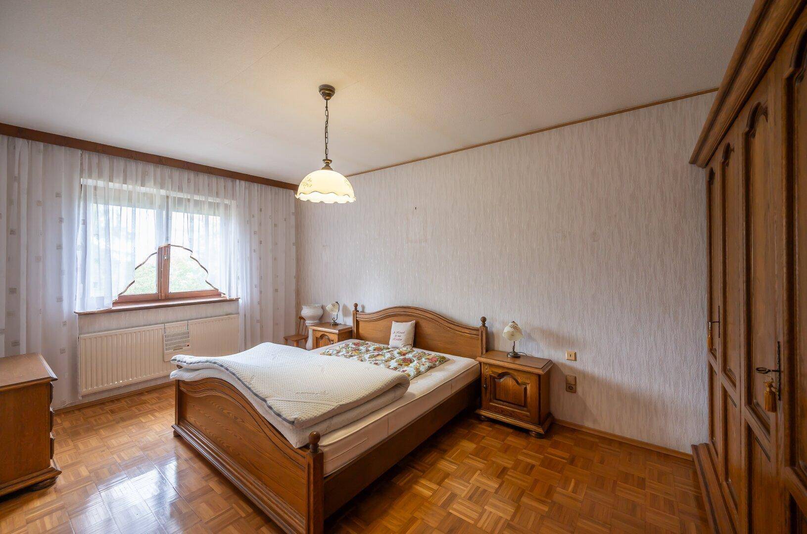 Schlafzimmer mit Holzmöbeln, Parkettboden und einem Fenster mit Vorhängen für natürliches Licht.