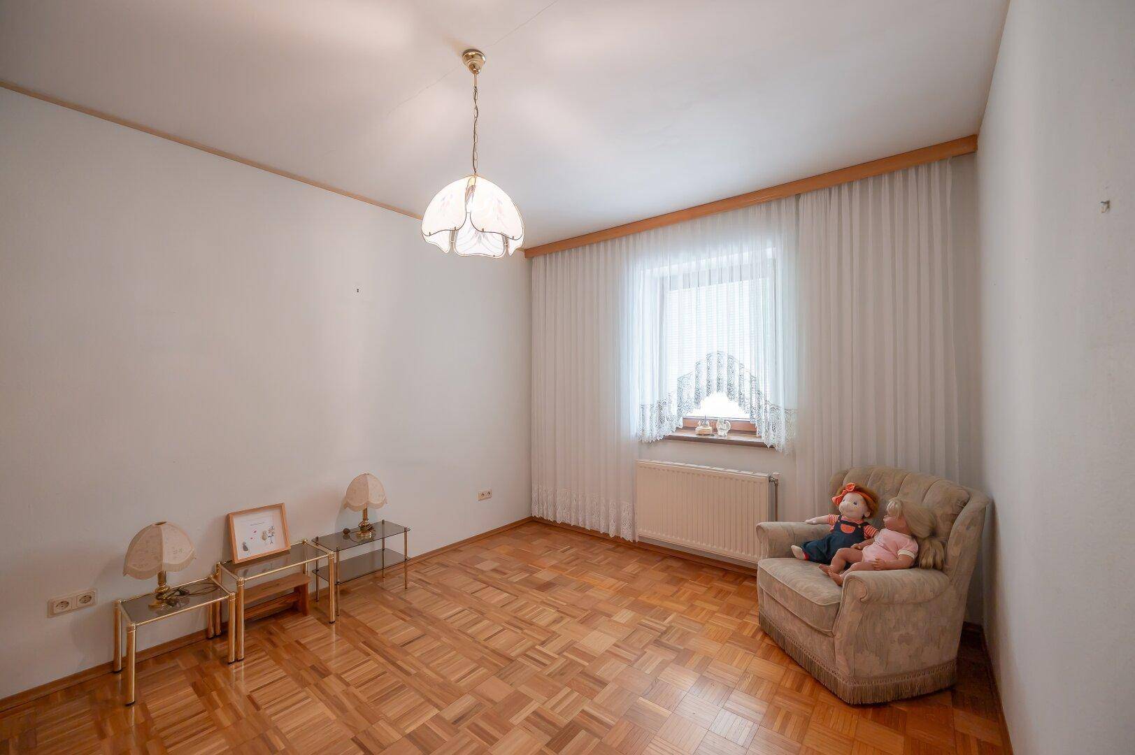Helles Zimmer mit Parkettboden, einem Sessel und einem Fenster mit Vorhängen.