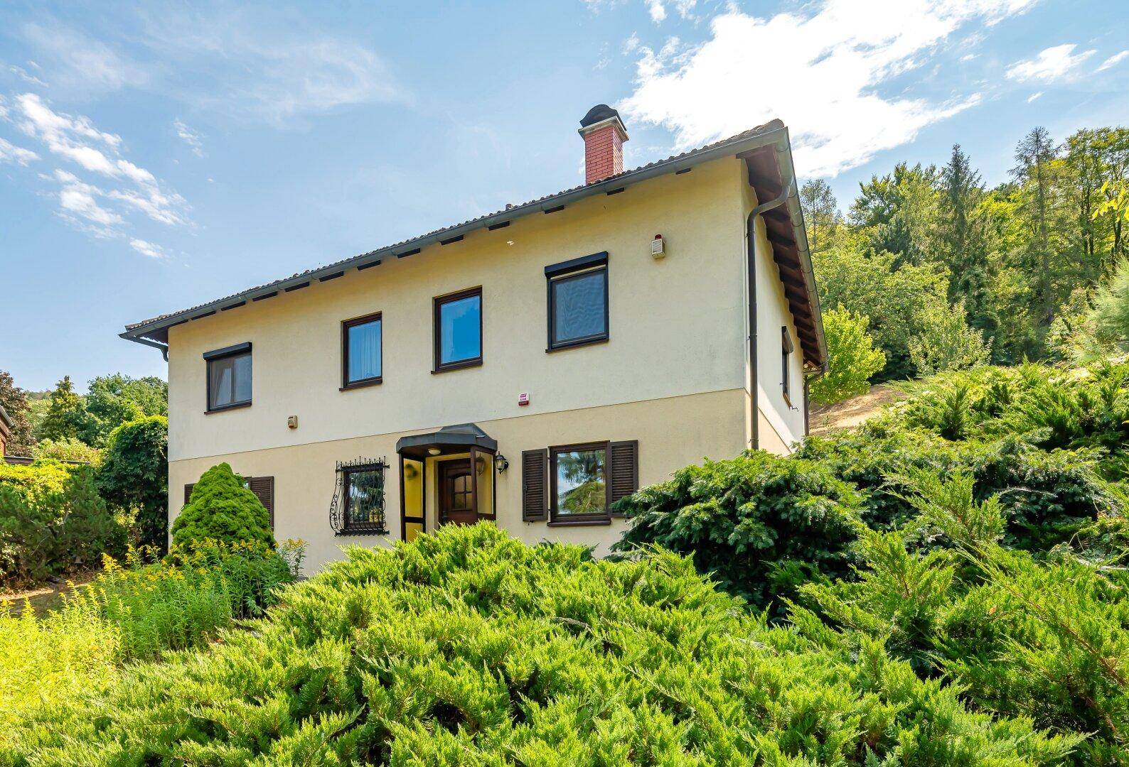 Ansicht des Hauses mit beiger Fassade, braunen Fensterläden und grünen Büschen im Vordergrund.