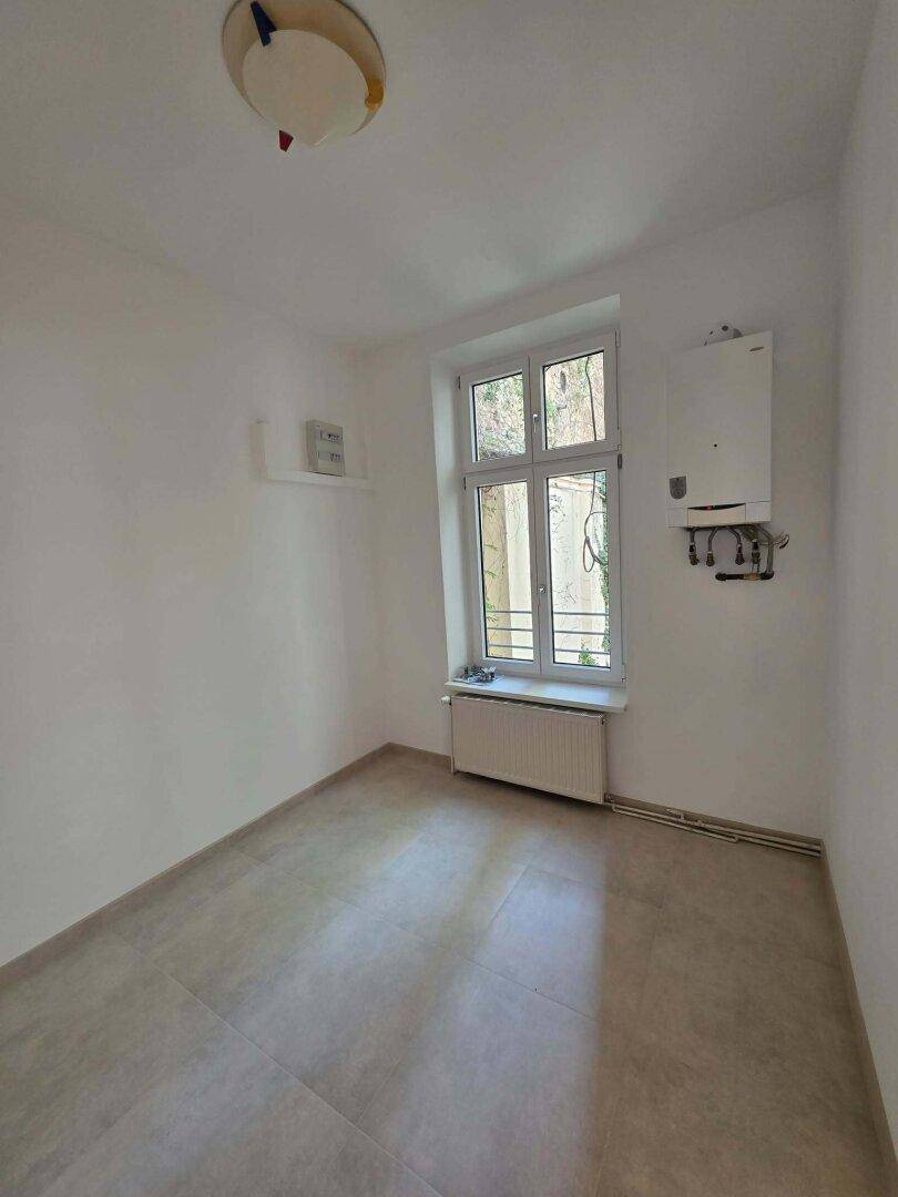 Kleiner Raum mit Fliesenboden, Fenster und einem an der Wand montierten Heizkessel.