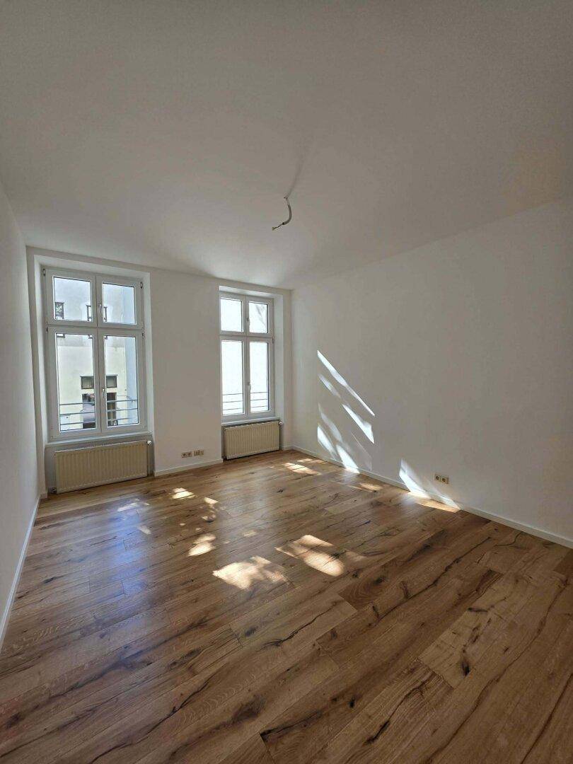 Leerer Raum mit Holzboden, zwei hohen Fenstern und weiß gestrichenen Wänden.