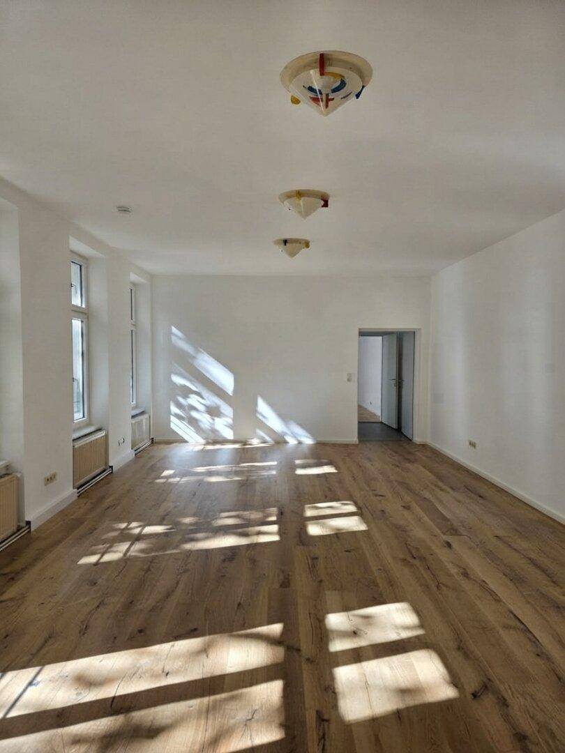 Langer, heller Wohnraum mit Holzboden, mehreren Fenstern und farbigen Deckenleuchten.