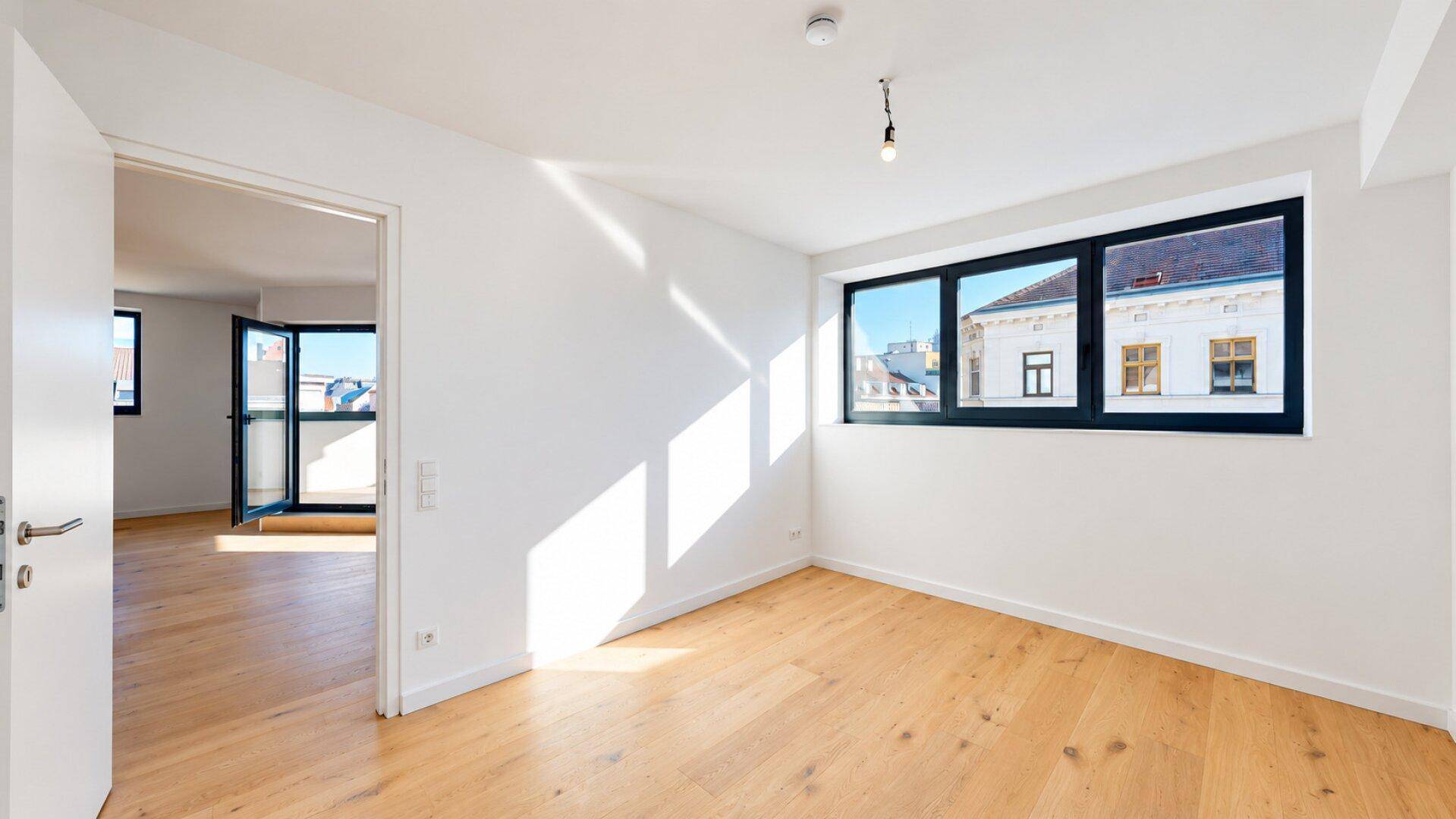 Helles Schlafzimmer mit Holzboden und Fenster, mit Blick in den angrenzenden Wohnbereich.