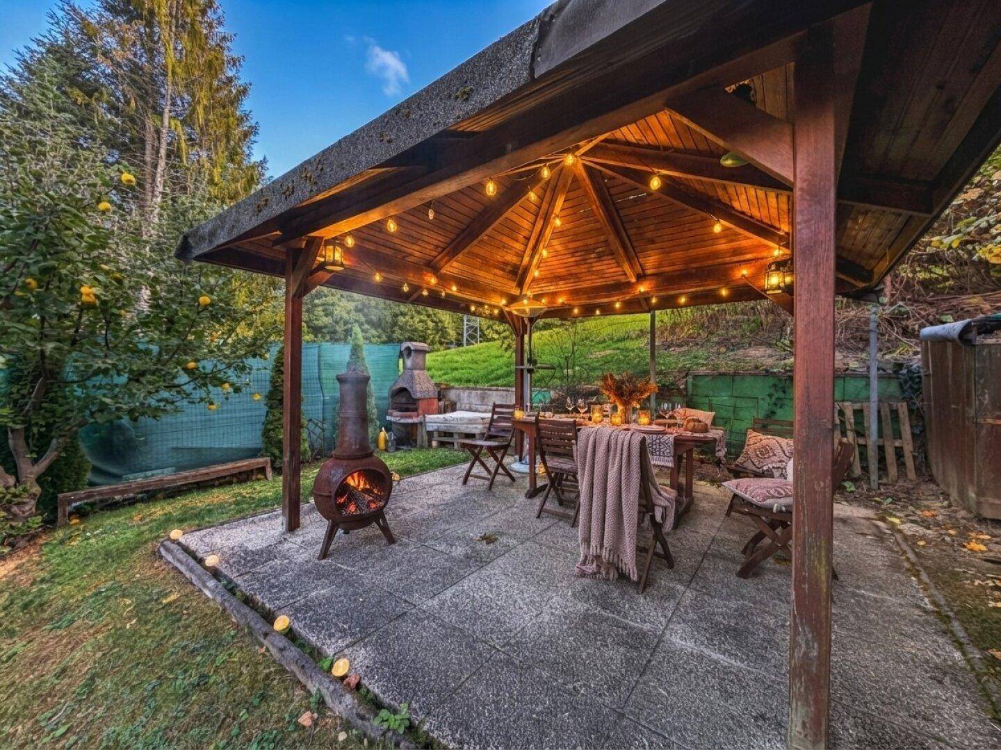 Stimmungsvolle überdachte Terrasse am Abend mit Lichterketten, Feuerstelle und Esstisch.