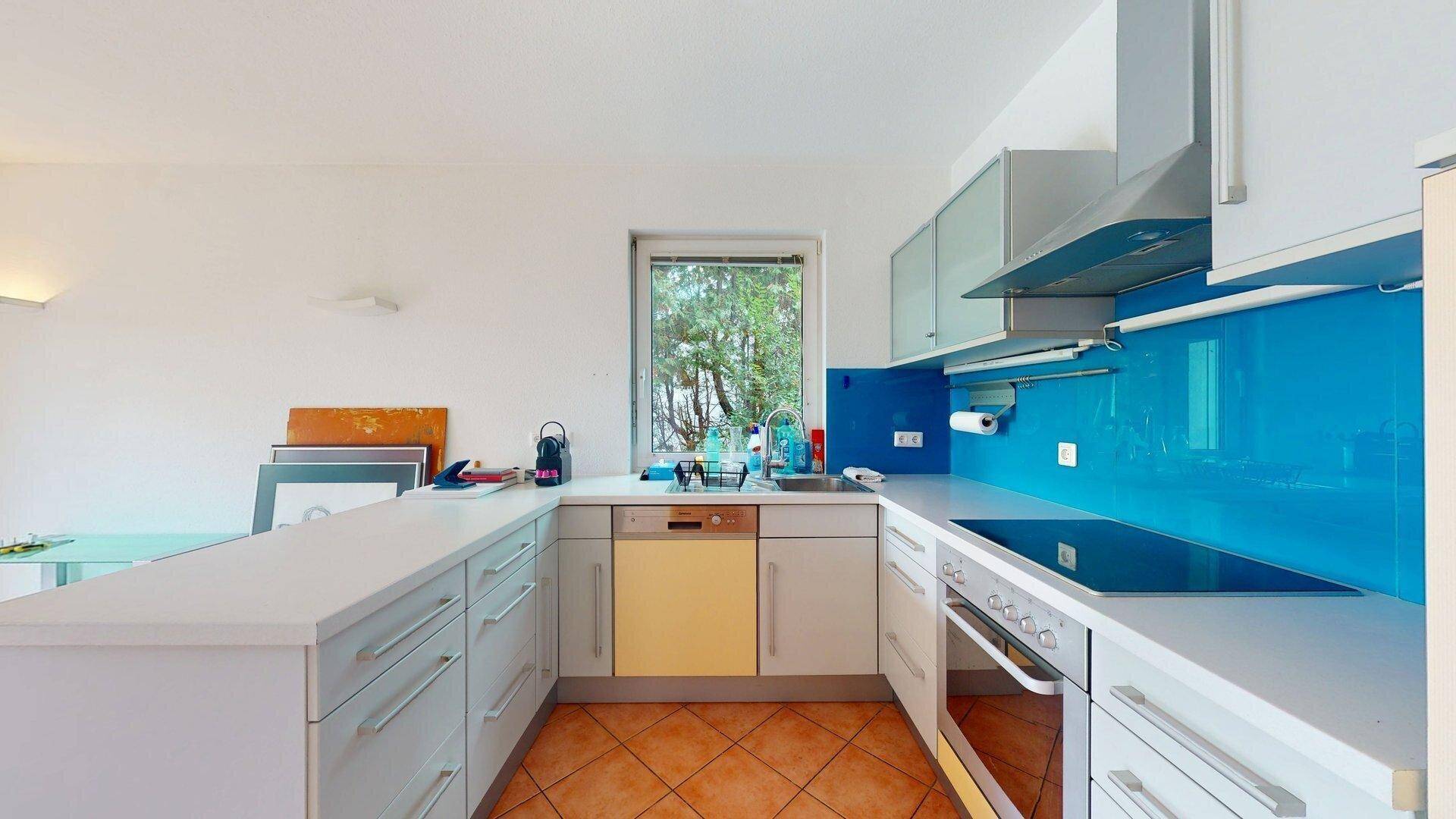 Moderne Küche mit weißen Schränken, blauer Rückwand, Fenster und integrierten Geräten.