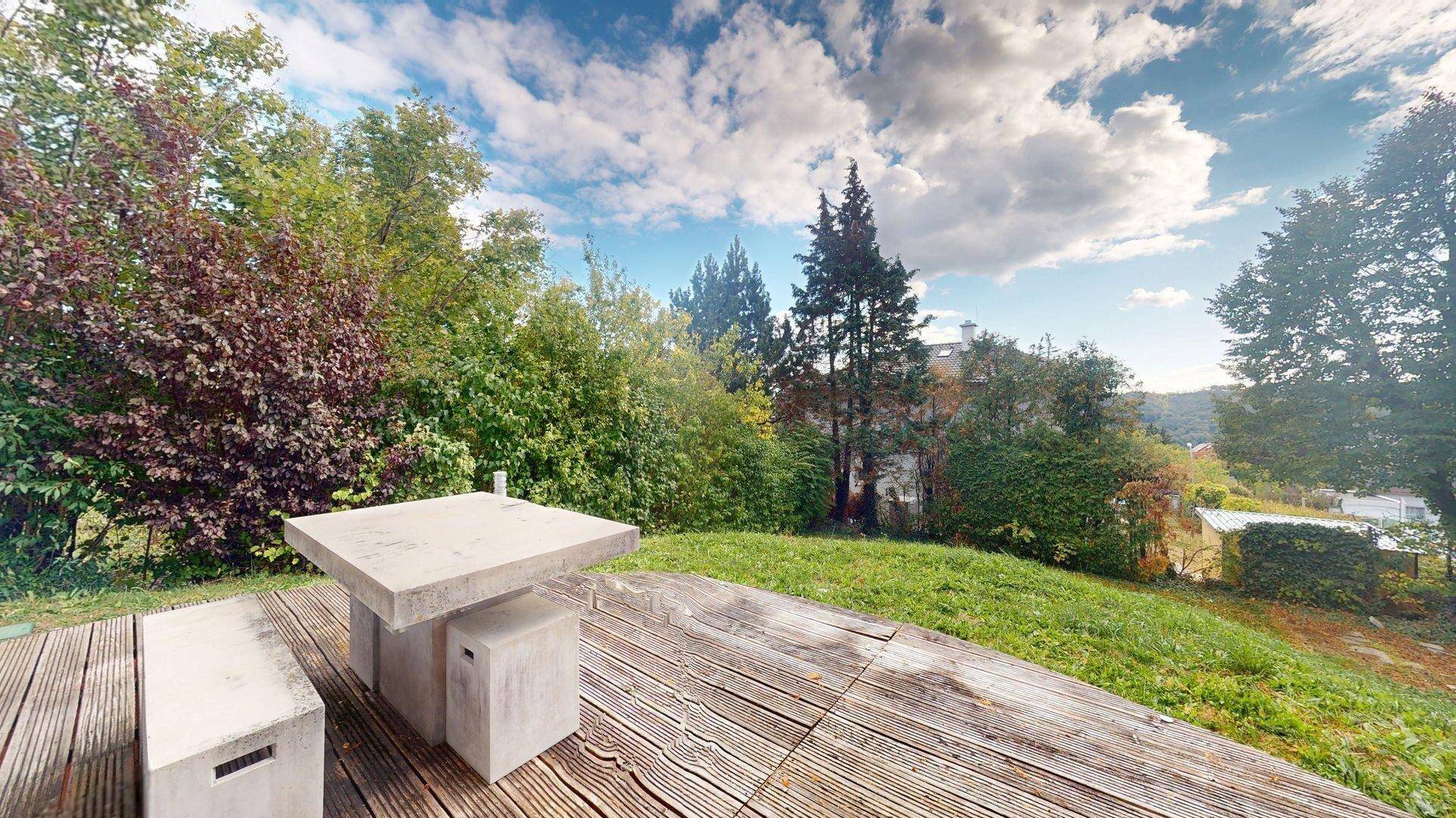 Holzterrasse mit Steintisch und Bänken, umgeben von viel Grün und einem weiten Blick in die Landschaft.